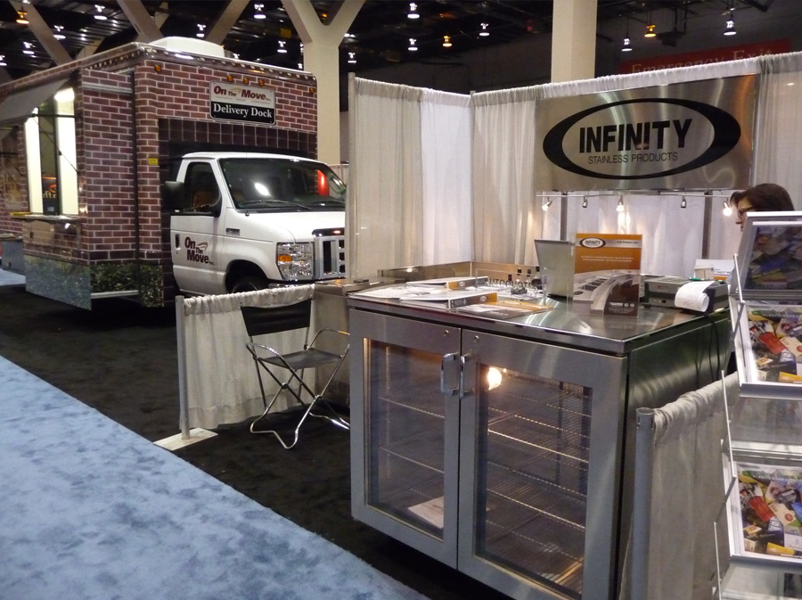 image of trade show booth with logo sign reading infinity stainless products and underbar refrigerator with glass doors and racks